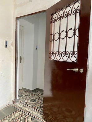 Una habitación con piso estampado, una puerta con reja metálica y una pared blanca con interruptor de luz.