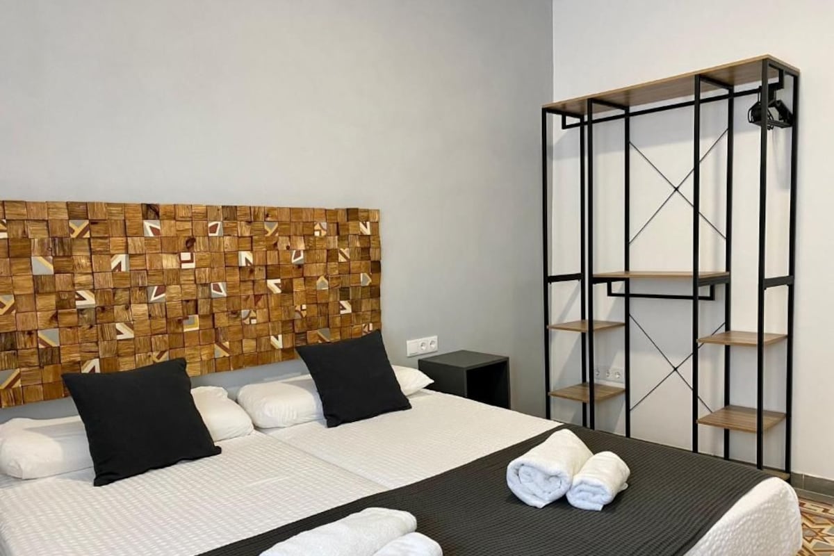 Et hotelværelse med en seng, en sengegavl med geometrisk mønster, en sort metalreol og et gulv med et mønster.