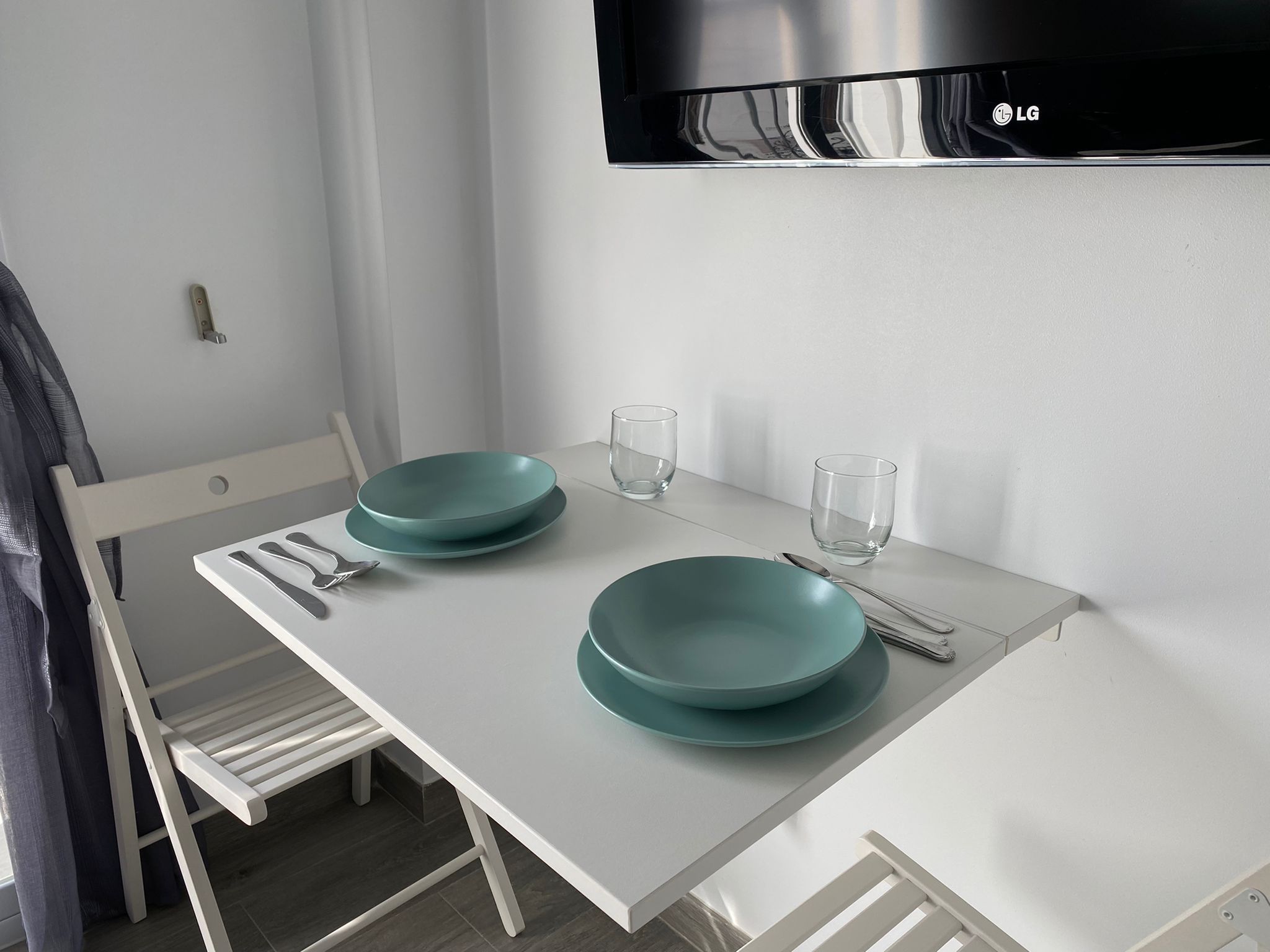 A white dining table with two teal plates and two clear glasses, a white chair, and a wall-mounted TV.