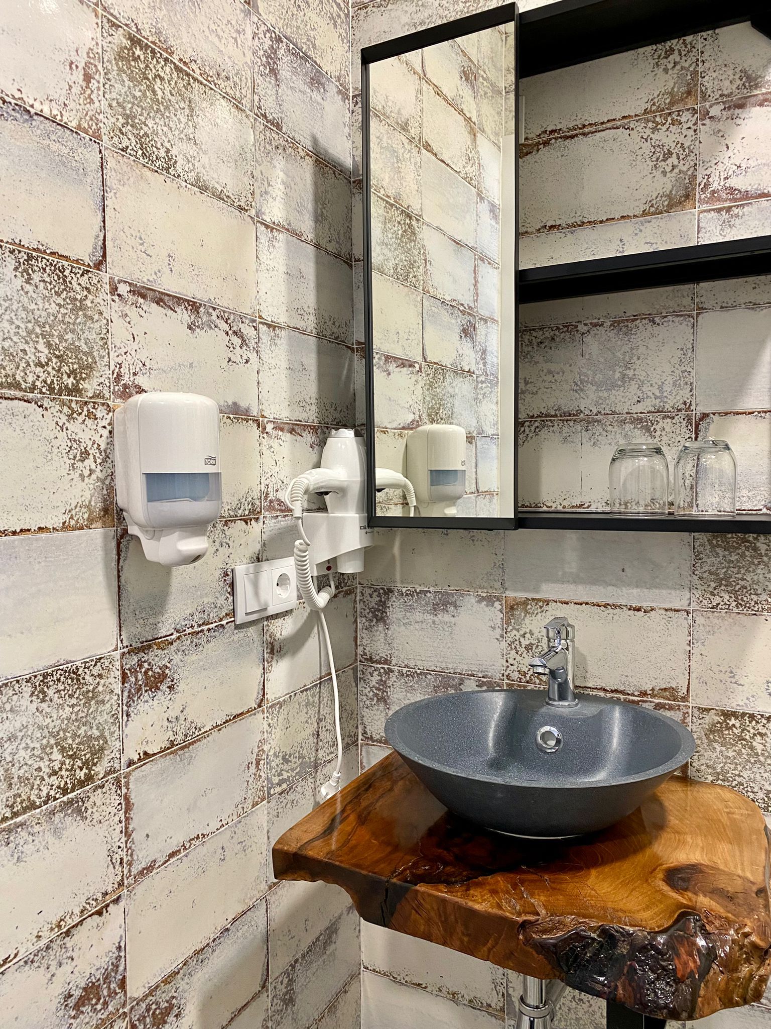 A bathroom with a rustic tiled wall, a wooden sink, a mirror, and a hand dryer.