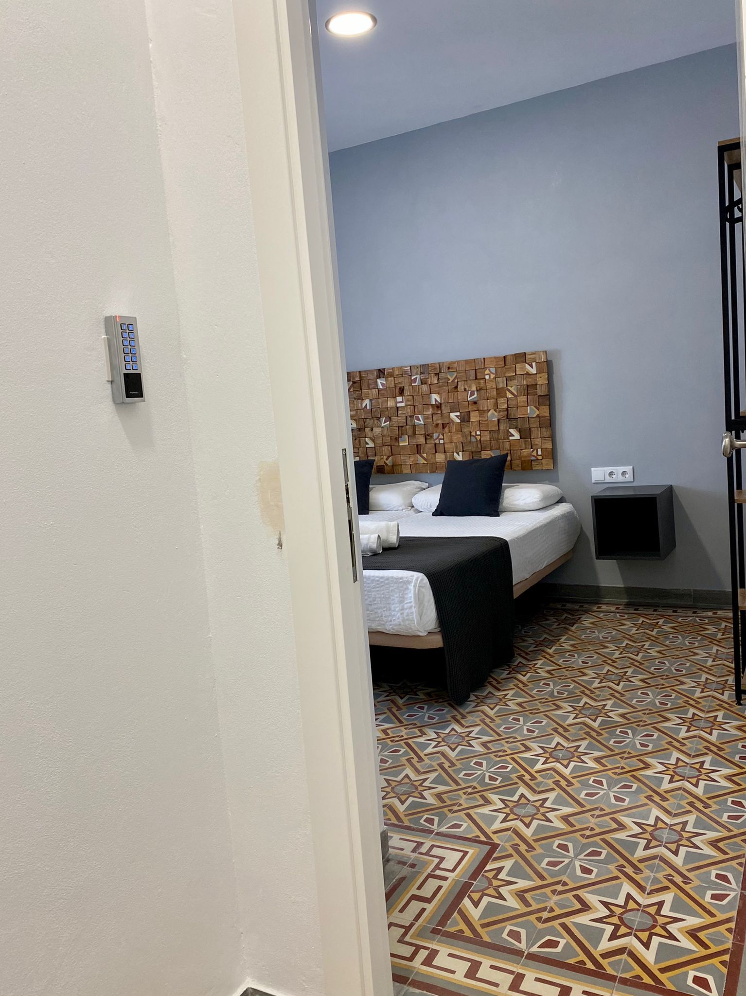 A hotel room with a patterned floor, a bed with white bedding and a dark headboard, and a wall-mounted control panel.