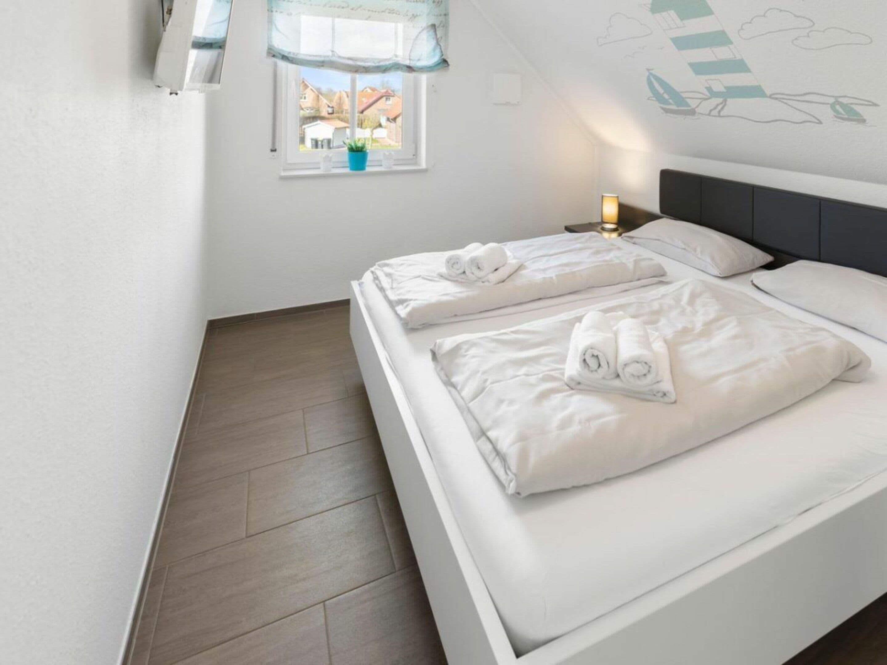 A hotel room with two beds, a window with a view of houses, a wall with a leaf pattern, and a bedside lamp.