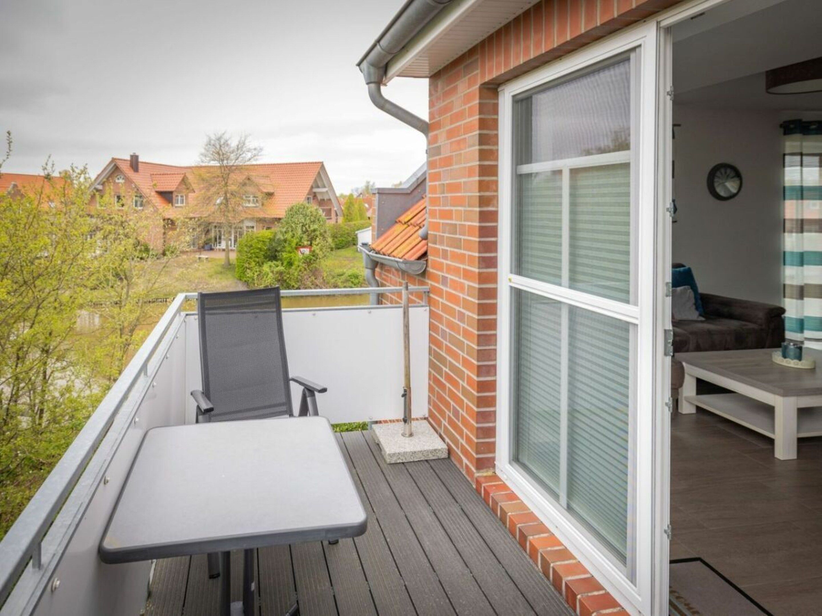 A balcony with a table and chair, a sliding glass door leading to an interior room with a sofa, and a view of neighboring houses.