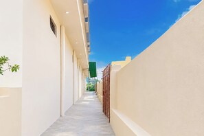Un estrecho pasillo con suelo de baldosas, bordeado por paredes lisas, que conduce a un edificio con entrada visible.