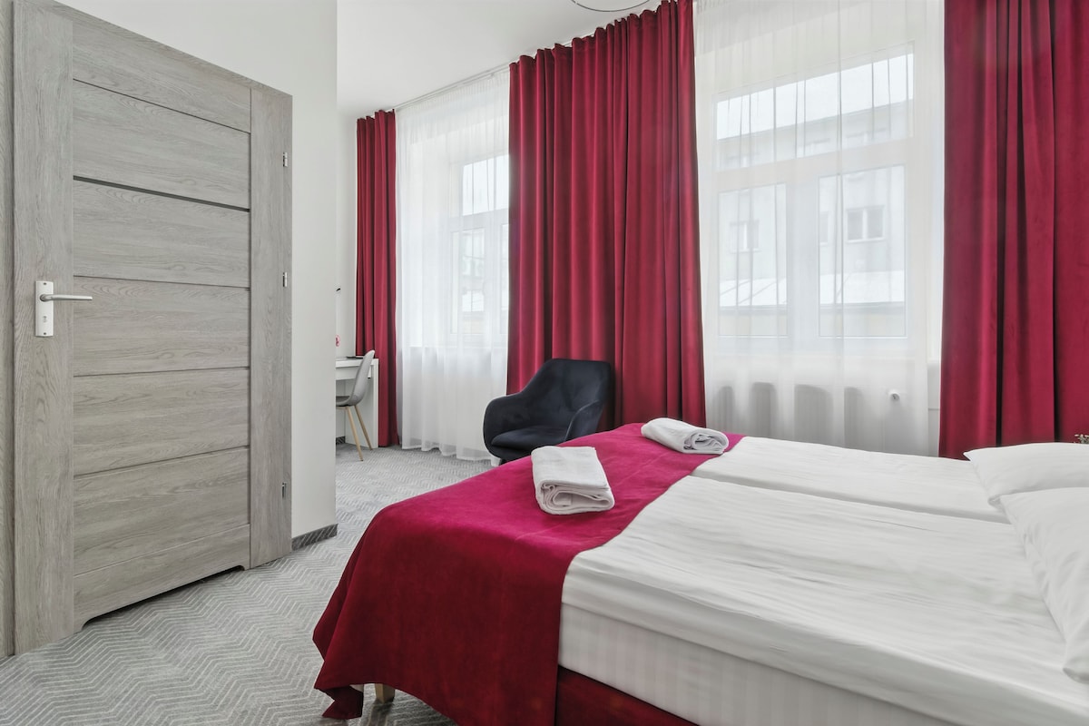 A hotel room with a wooden door, a desk with a chair, a bed with white linens, and red curtains.