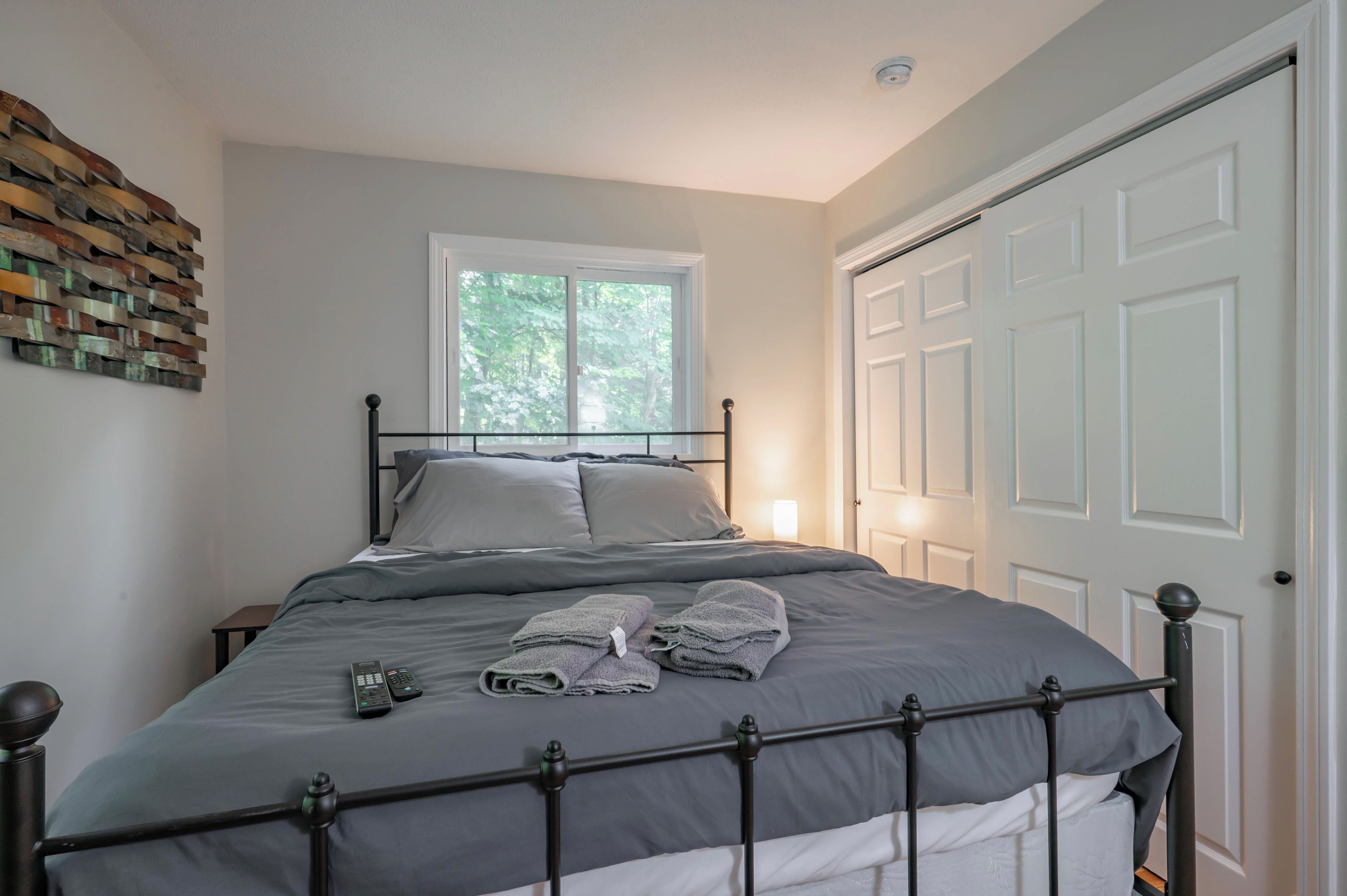 A neatly made bed with a metal frame, a window with a view of greenery, and a white door.