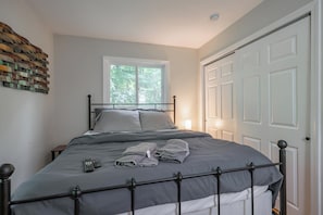 Ein ordentlich bezogenes Bett mit Metallgestell, ein Fenster mit Blick auf Grünflächen und eine weiße Tür.