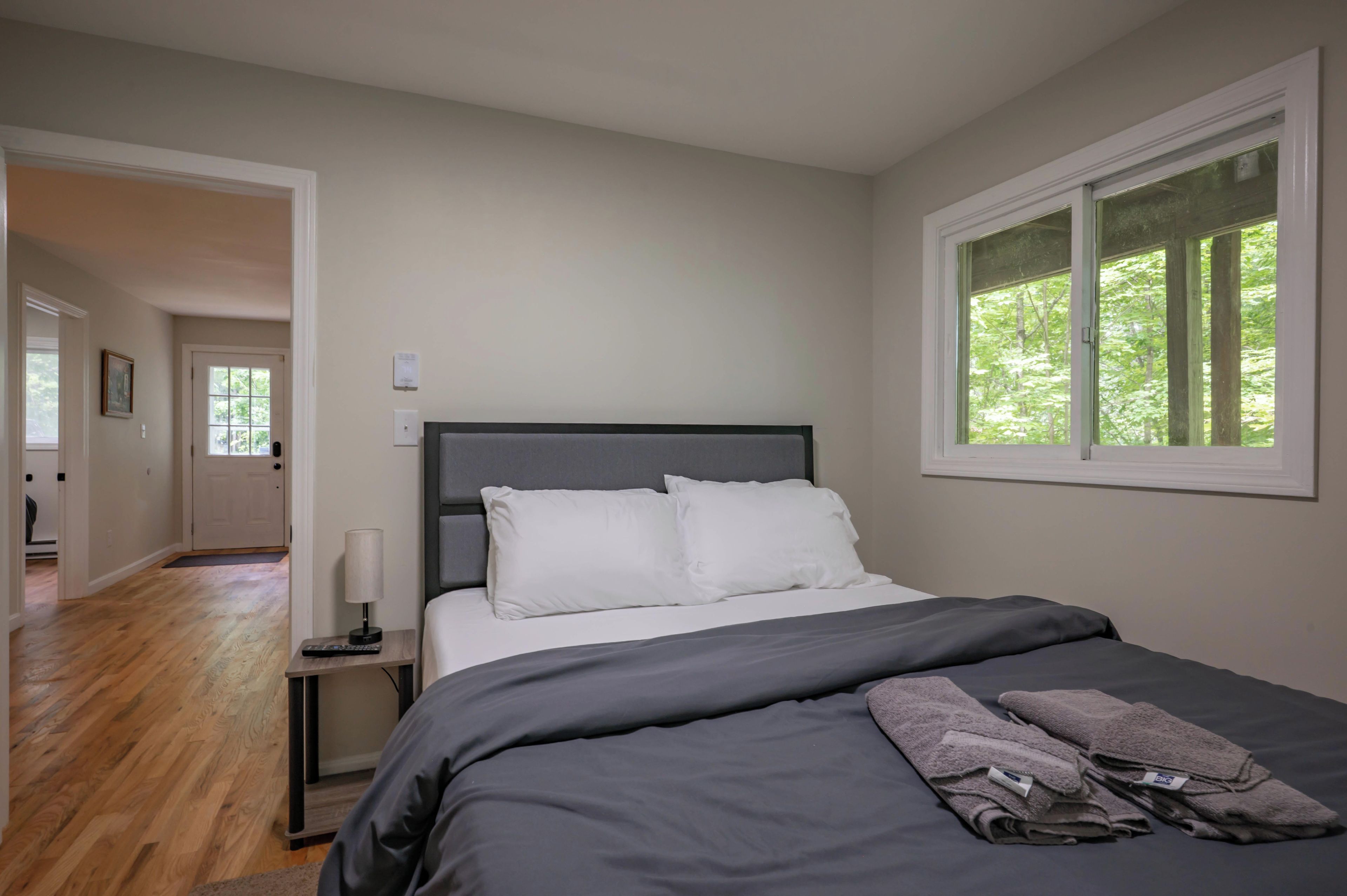 A bedroom with a bed, a nightstand, a lamp, a window with a view of trees, and a door leading to another room.