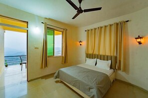 Une chambre avec un lit, un ventilateur de plafond, deux lampes fixées au mur, une fenêtre avec des rideaux et une vue sur l’extérieur.