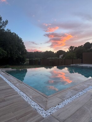 Une piscine rectangulaire dotée d’une terrasse en bois et bordée de galets, entourée d’un mur de briques et d’arbres.
