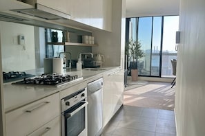 A modern kitchen with a gas stove, oven, and dishwasher. There is a large window providing a view of the city. - Luxury High Rise Suite & CBD Open Views. (Melbourne)