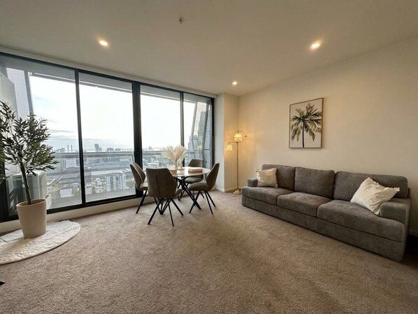 A modern living room with a large window, a sofa, a dining table with chairs, and a potted plant. - Luxury High Rise Suite & CBD Open Views. (Melbourne)