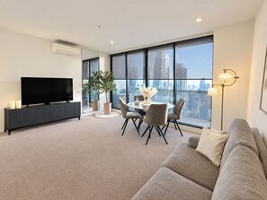 A modern living room with a flat-screen TV, a dining table with chairs, a sofa, a floor lamp, and large windows offering a city view. - Luxury High Rise Suite & CBD Open Views. (Melbourne)