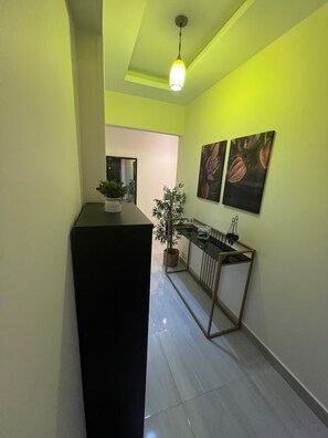 A hallway with a black reception desk, a potted plant, a console table with decorative items, and two wall-mounted paintings. - Central Mokattam Apt – 2BR w/ AC, Near Everything (Cairo Governorate)