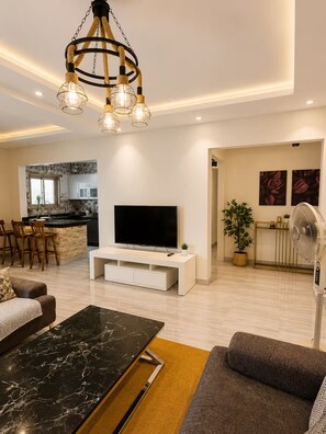 A modern living room with a flat-screen TV, a marble coffee table, a sofa, and a dining area with a kitchen in the background. - Central Mokattam Apt – 2BR w/ AC, Near Everything (Cairo Governorate)