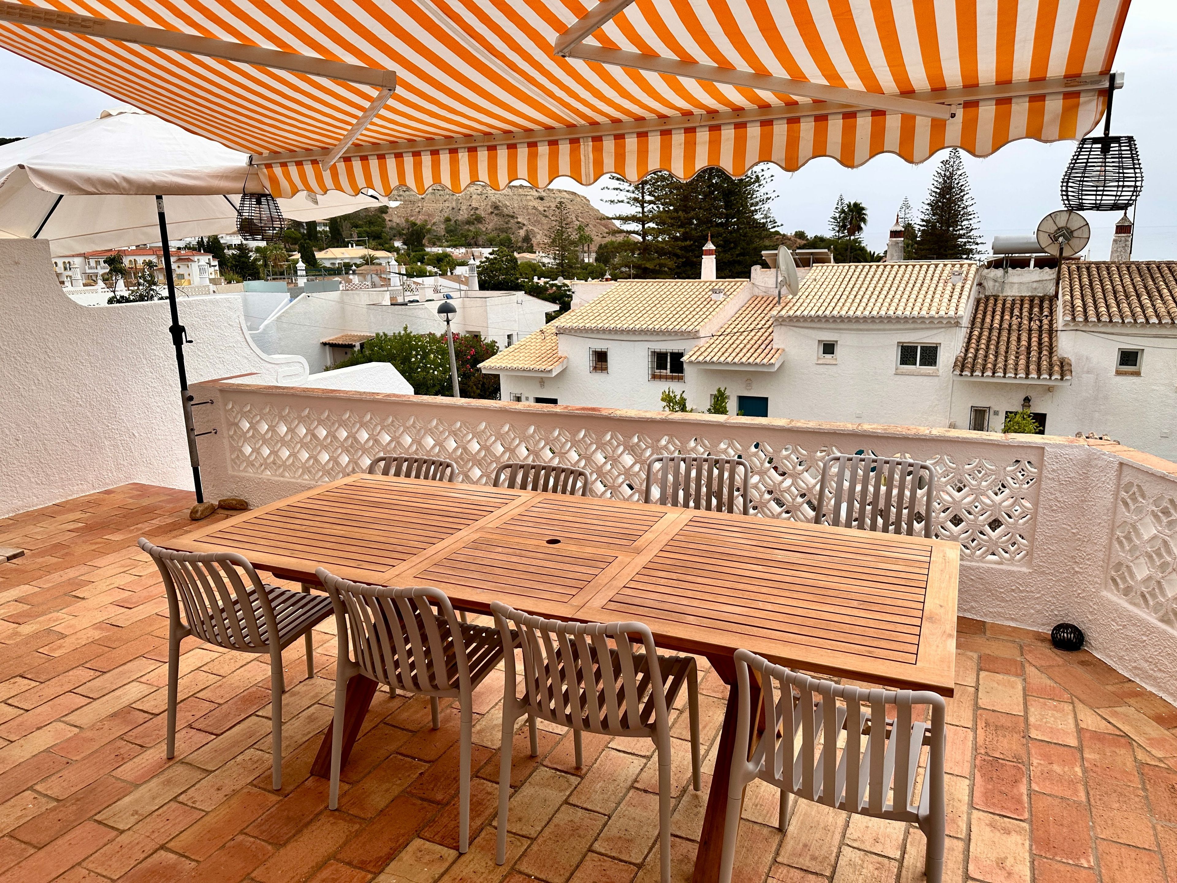 Una terraza con mesa y sillas de madera, un toldo a rayas y vistas de edificios al fondo.