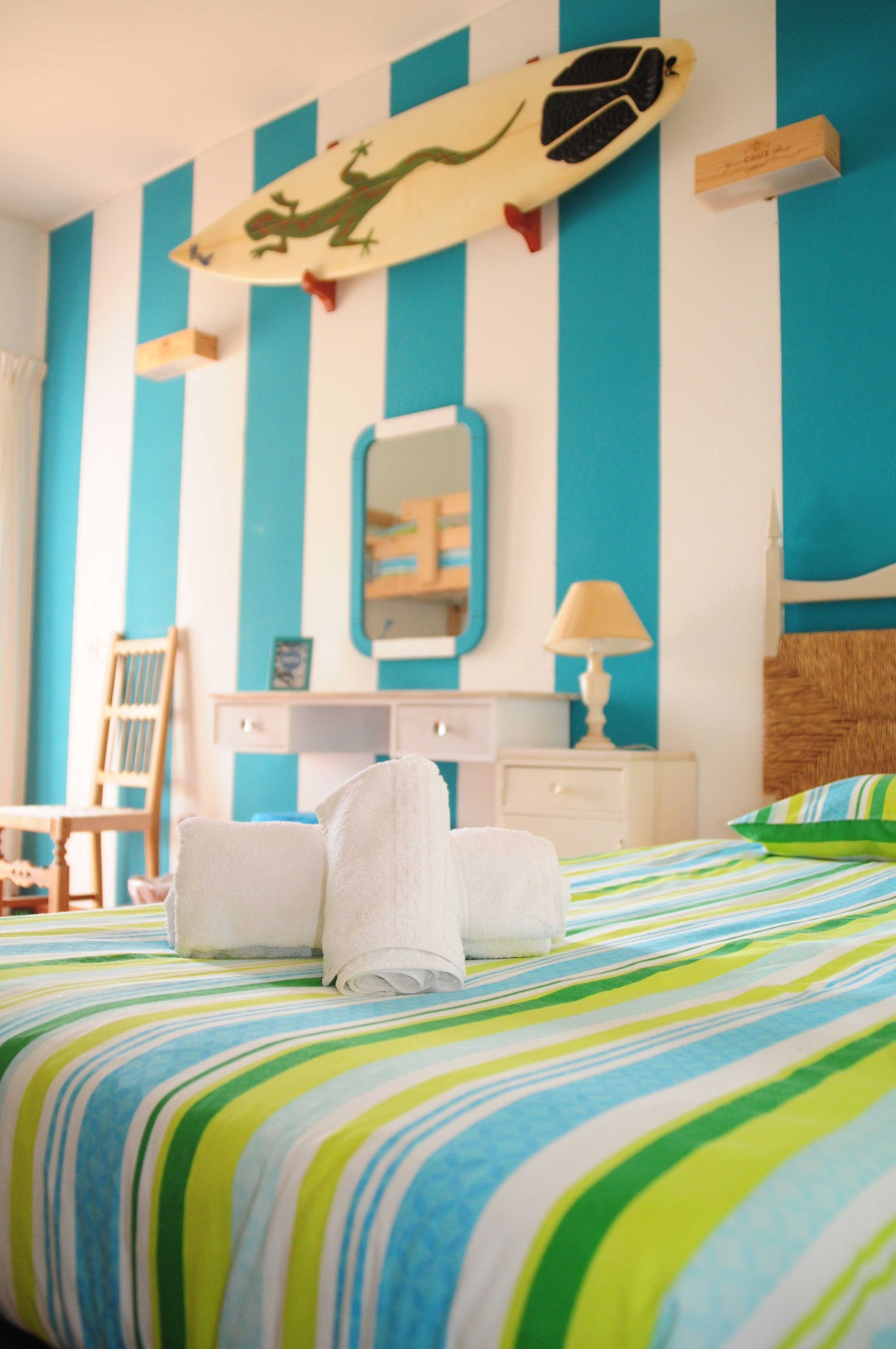 A hotel room with a bed, a chair, a desk, a mirror, and a surfboard decoration on the wall.