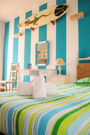 Une chambre d’hôtel avec un lit, une chaise, un bureau, un miroir et une décoration murale représentant une planche de surf.