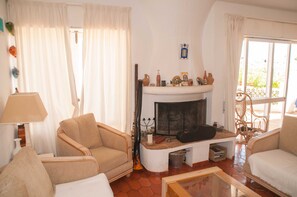 Un salon avec une cheminée, deux fauteuils, une lampe et une fenêtre avec des rideaux.
