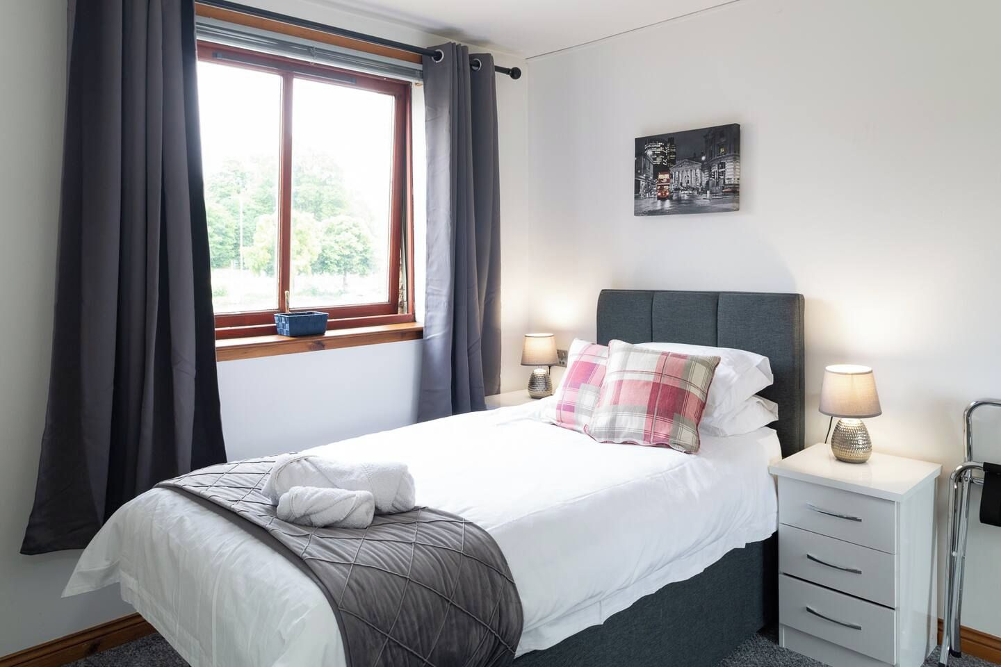 A neatly made bed with a pillow, a bedside table with a lamp, and a window with curtains.
