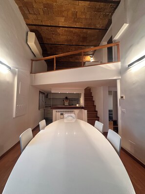Uma sala de jantar espaçosa com uma mesa comprida e cadeiras, cozinha ao fundo e escada que dá para um andar superior.