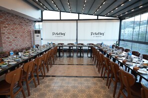 Un salón de conferencias con una larga mesa dispuesta para una comida, rodeada de sillas, con una pared de ladrillo y ventanas amplias en un lateral.