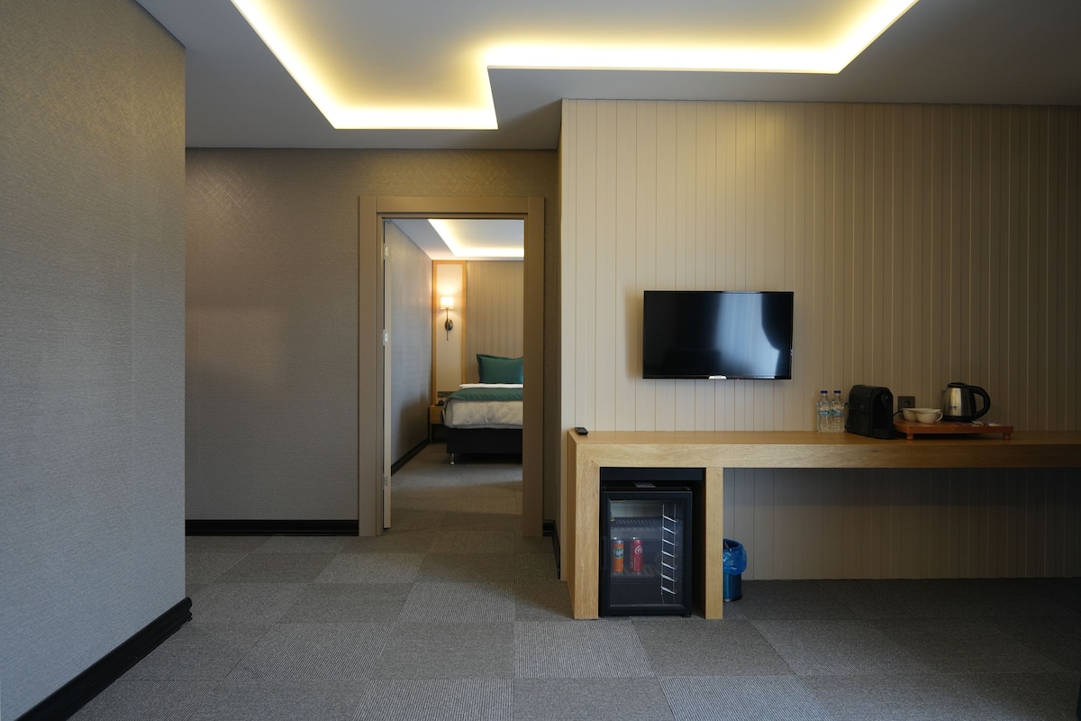 Habitación de hotel con televisor de pantalla plana en la pared, una kitchenette pequeña con cafetera y refrigerador minibar.