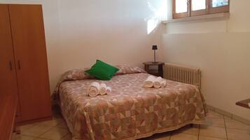 Ein Schlafzimmer mit einem Bett, einem grünen Kopfkissen, zusammengerollten Handtüchern, einem Nachttisch mit Lampe, einem Heizkörper und einem Holzschrank.