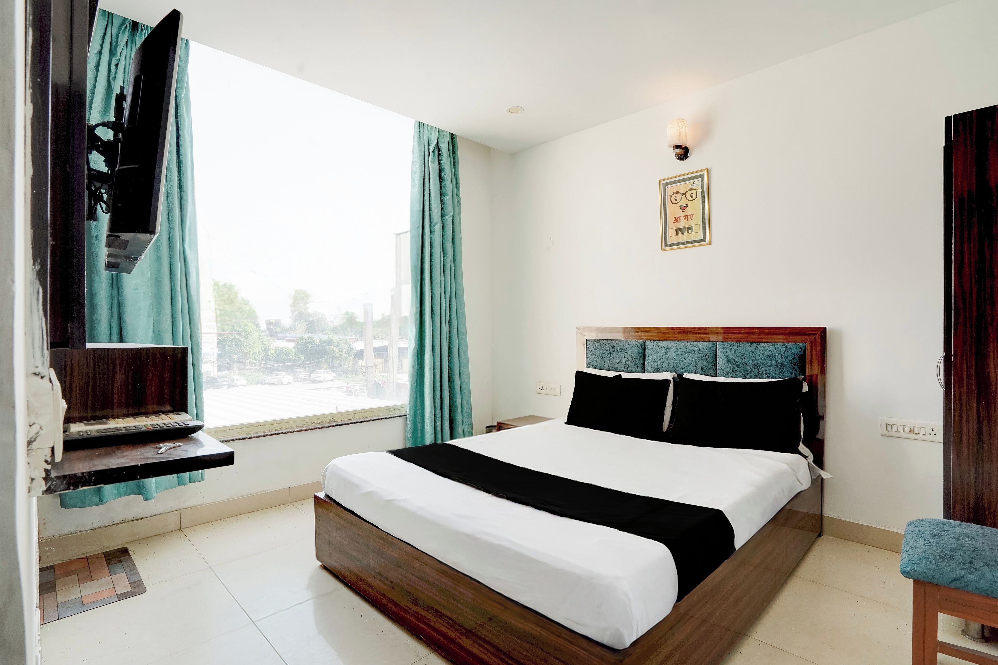 A hotel room with a bed, a desk with a computer, a chair, a window with curtains, and a wall-mounted light fixture.