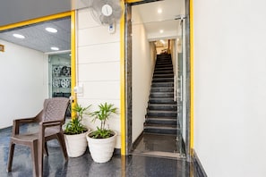 Un área de recepción con una silla, dos plantas en macetas, un ventilador y una escalera que sube a un piso superior.