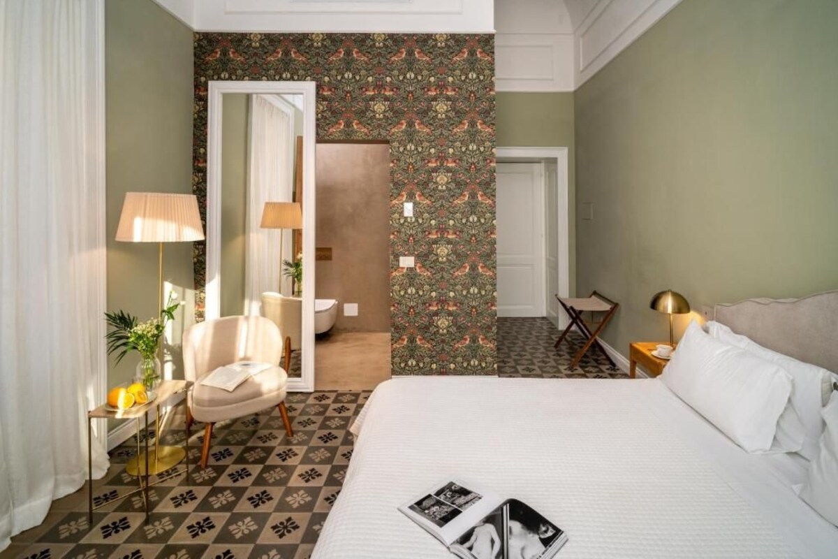 A hotel room with a bed, a chair, a small table, a lamp, and a mirror.