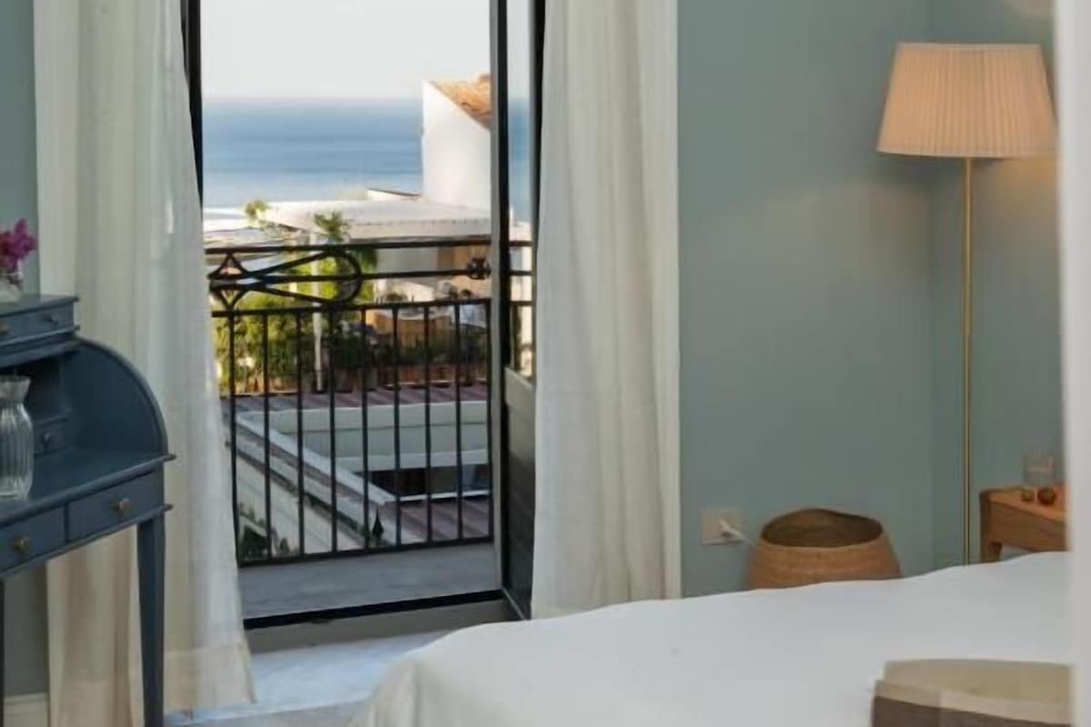 A bedroom with a bed, a nightstand, a lamp, and a view of the sea through an open door.