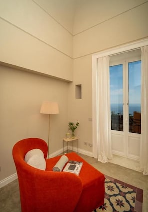 A room with a red chair, a small table with a lamp, and a view through a window with curtains. - Villa Mabel Boutique Hotel (Taormina)