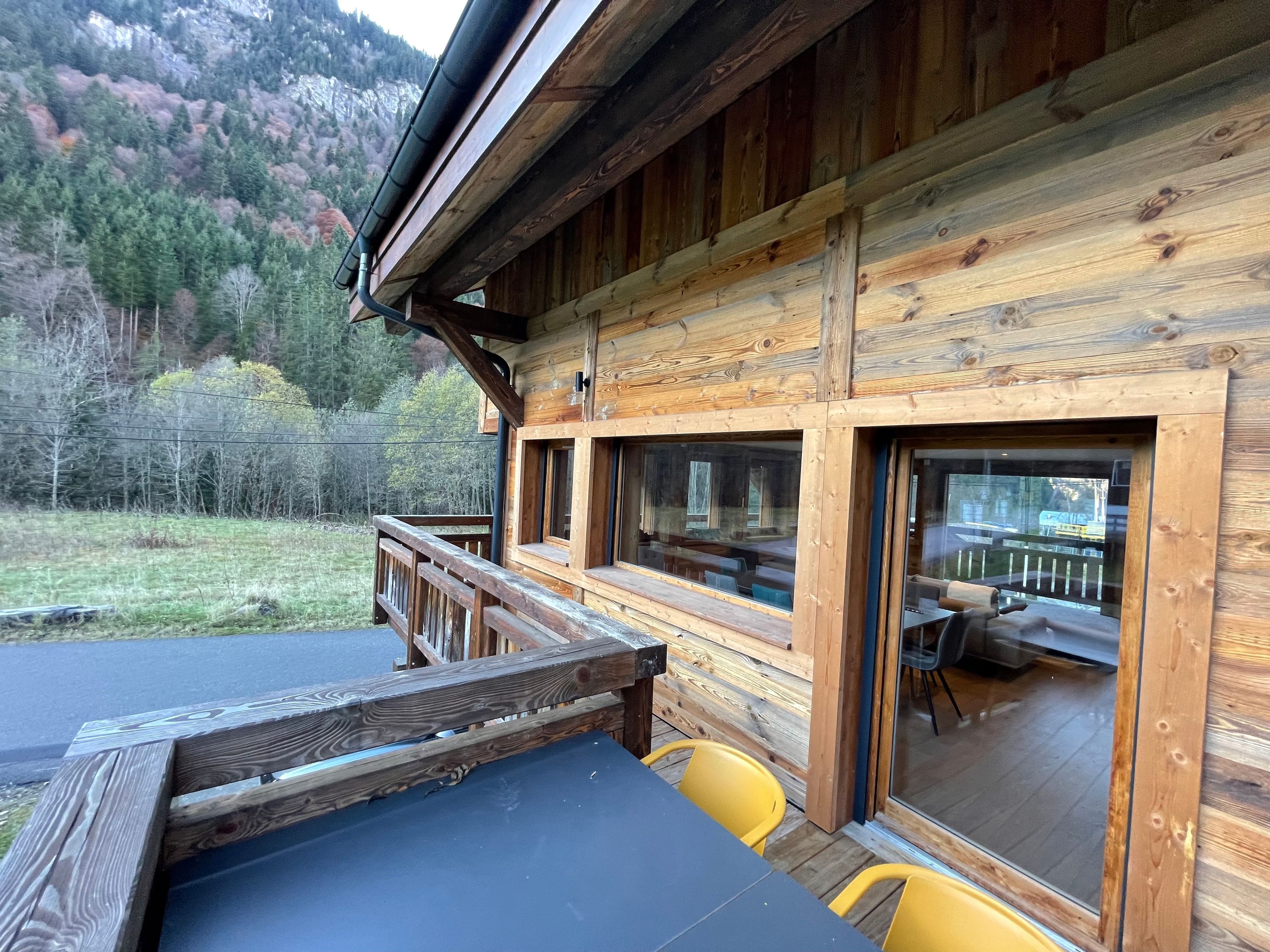 Eine Holzhütte mit einer Terrasse, Sitzgelegenheiten im Freien und Blick auf einen bewaldeten Berg.
