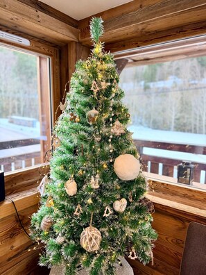 Un sapin de Noël décoré d’ornements et illuminé, installé à l’intérieur d’une cabane aux murs et aux fenêtres en bois.