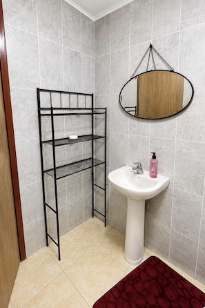 A bathroom with a white pedestal sink, a black metal shelf unit, and a round mirror with a wooden frame. - Moorish Mount Cool Breeze Room – Bright Hilltop Double, Mandeville (Mandeville)