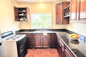 A kitchen with dark wooden cabinets, a gas stove, a sink, and a window with a view of greenery. - Moorish Mount Cool Breeze Room – Bright Hilltop Double, Mandeville (Mandeville)