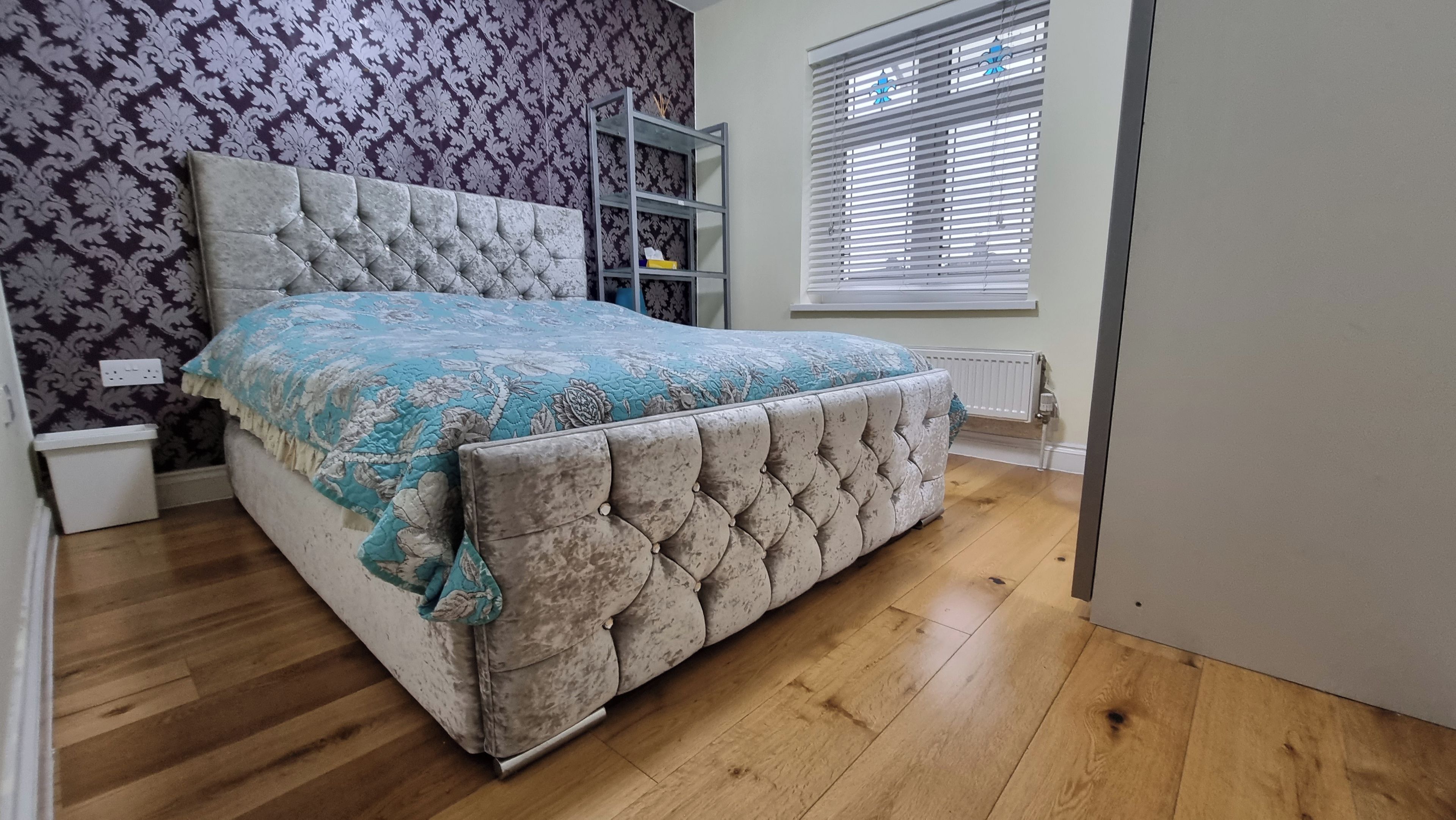 Une chambre à coucher avec une tête de lit capitonnée, une couvre-lit à motifs floraux et une fenêtre avec des stores.