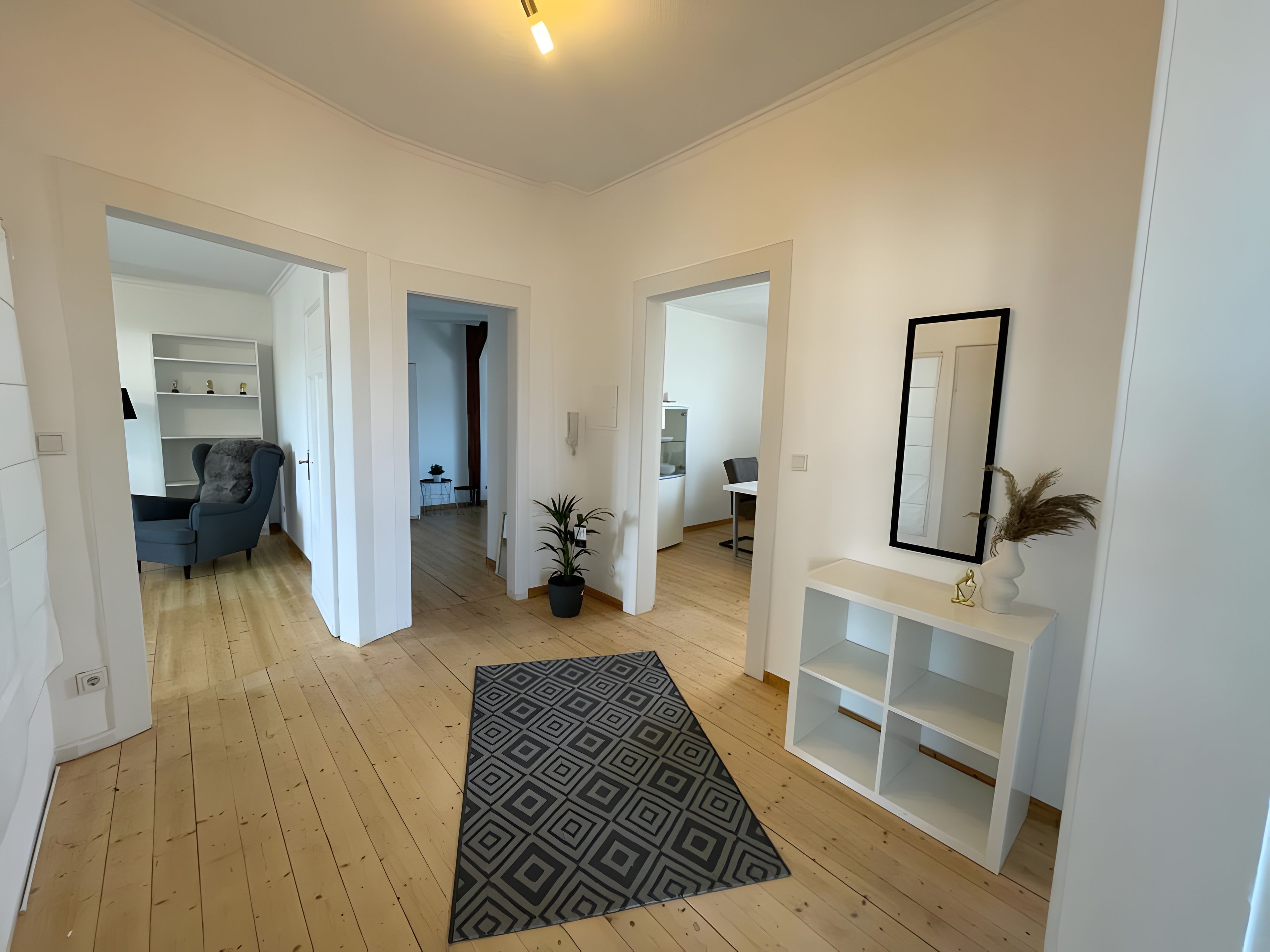 Un hall d’entrée spacieux, doté d’un parquet en bois, d’un tapis à motifs et d’une console blanche agrémentée d’un miroir et d’un vase.
