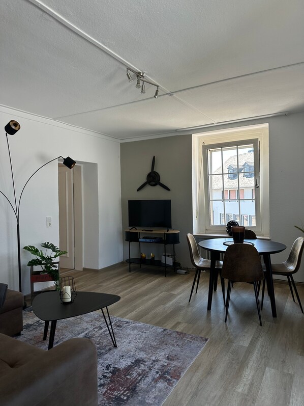 Un salon moderne doté d’un téléviseur à écran plat, d’une table à manger avec des chaises, d’un lampadaire et d’une grande fenêtre offrant une vue sur les bâtiments.