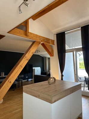 Ein modernes Interieur mit freiliegenden Holzbalken, einer zentralen Kochinsel mit Ablagefläche und Blick ins Freie.