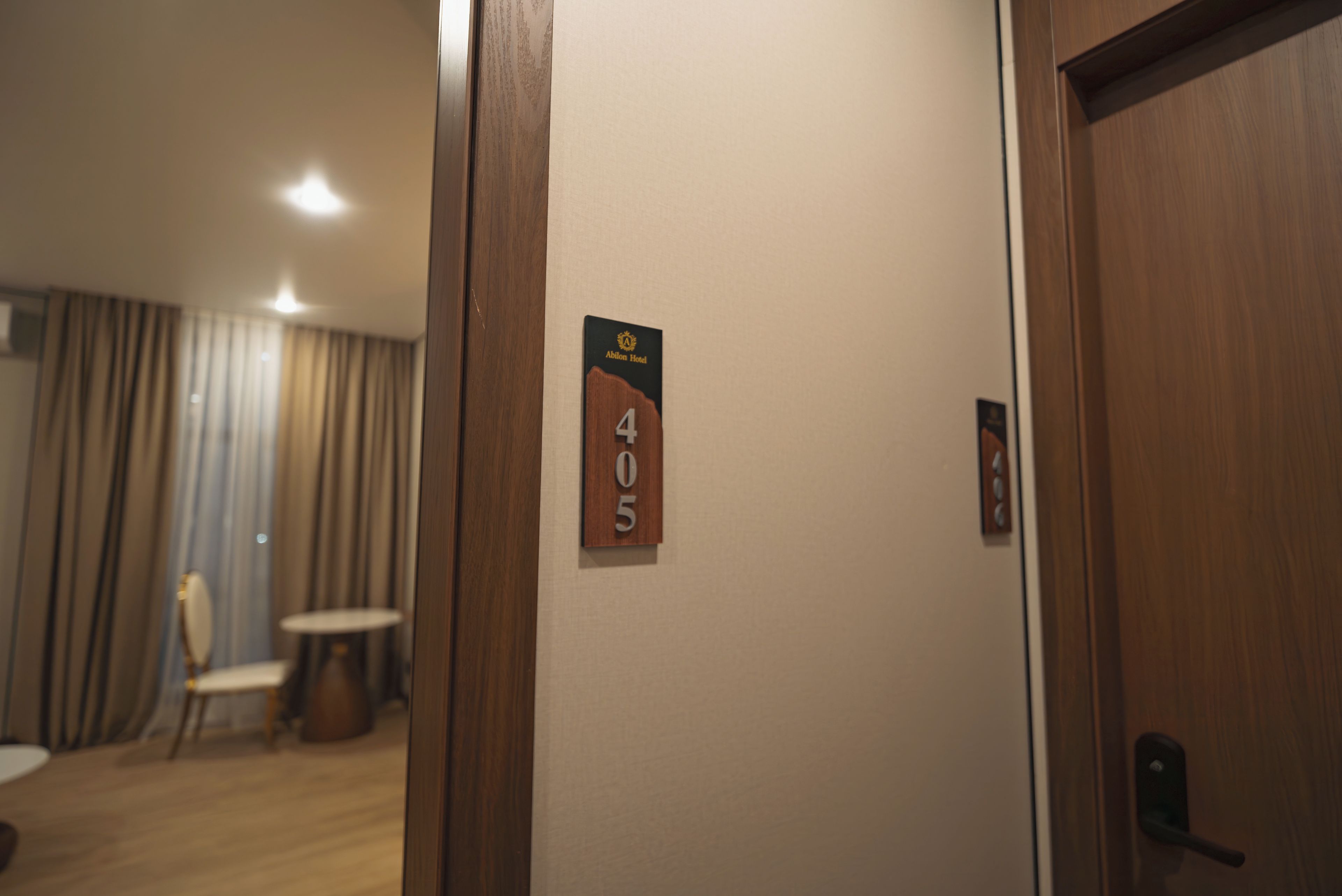 Habitación de hotel con puerta de madera, un cartel con número en la pared y una vista parcial de una habitación con una silla y una mesa.