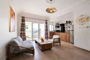 A modern living room with a sofa, a wooden coffee table, and a chair. There is a kitchen area with a stove, a refrigerator, and a microwave. The room has a large window with curtains and a ceiling light. - New Listing (Yaoundé)