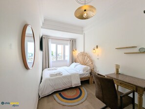 Habitación de hotel con cama, escritorio, silla, espejo redondo, lámpara en el techo y ventana con cortinas.