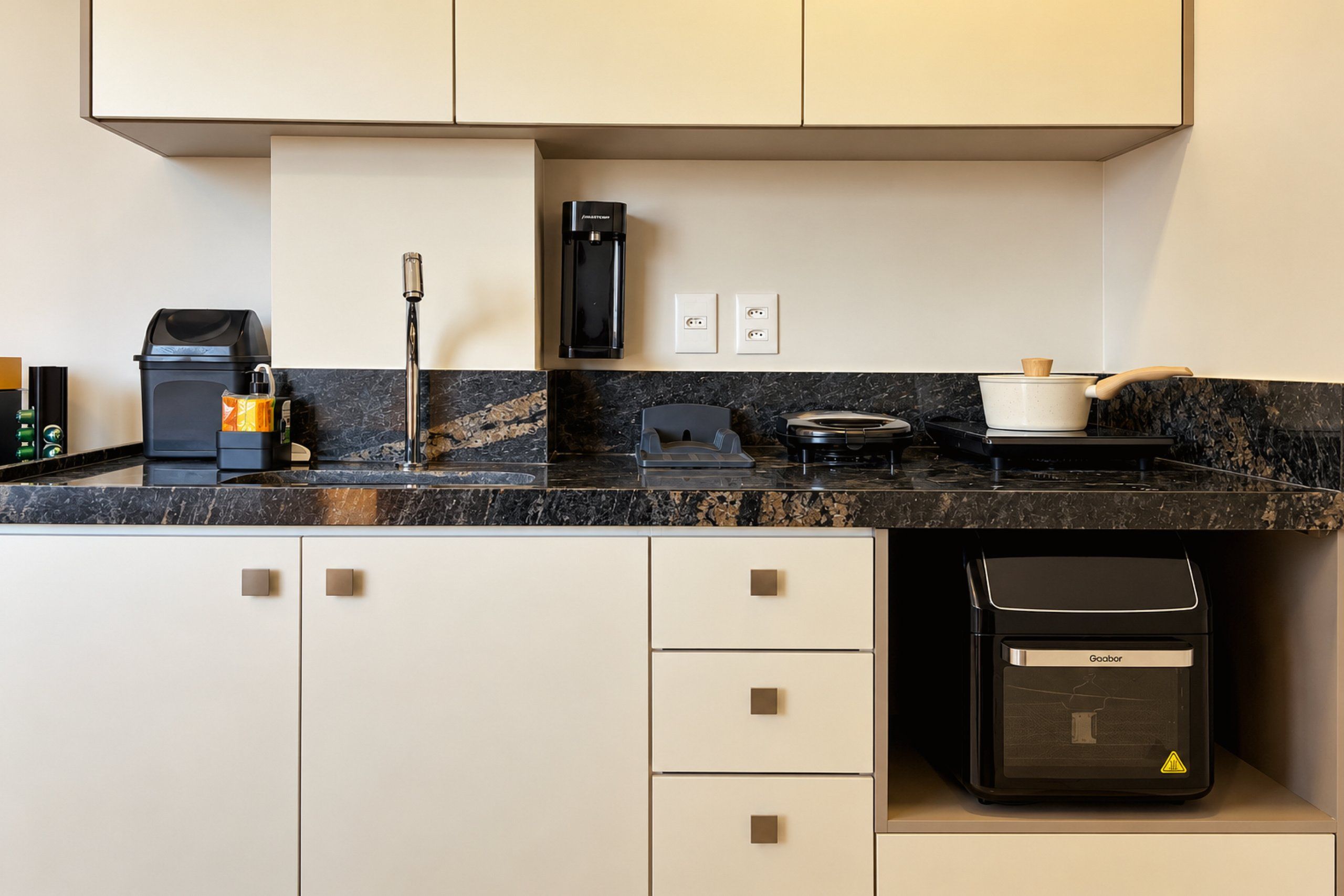 Una cocina moderna con encimera negra, armarios blancos, máquina de café, hervidor y cocedor de arroz.