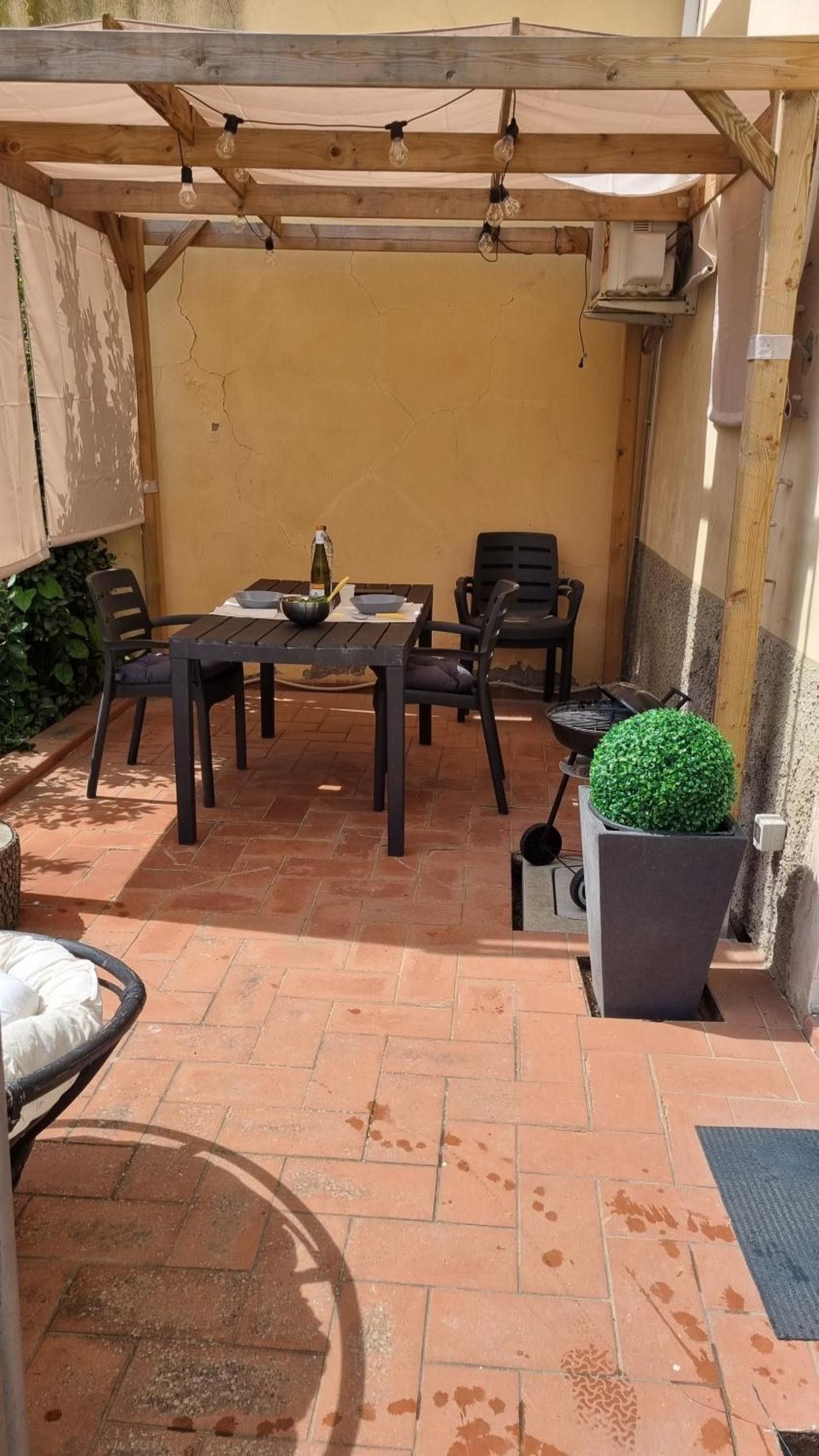 Un espace extérieur aménagé en terrasse, avec une table et des chaises, un pot de fleurs et une pergola illuminée de guirlandes lumineuses.