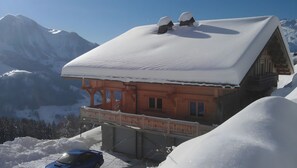 Una xalet amb teulada coberta de neu, balcó de fusta i un paisatge de muntanya al fons.
