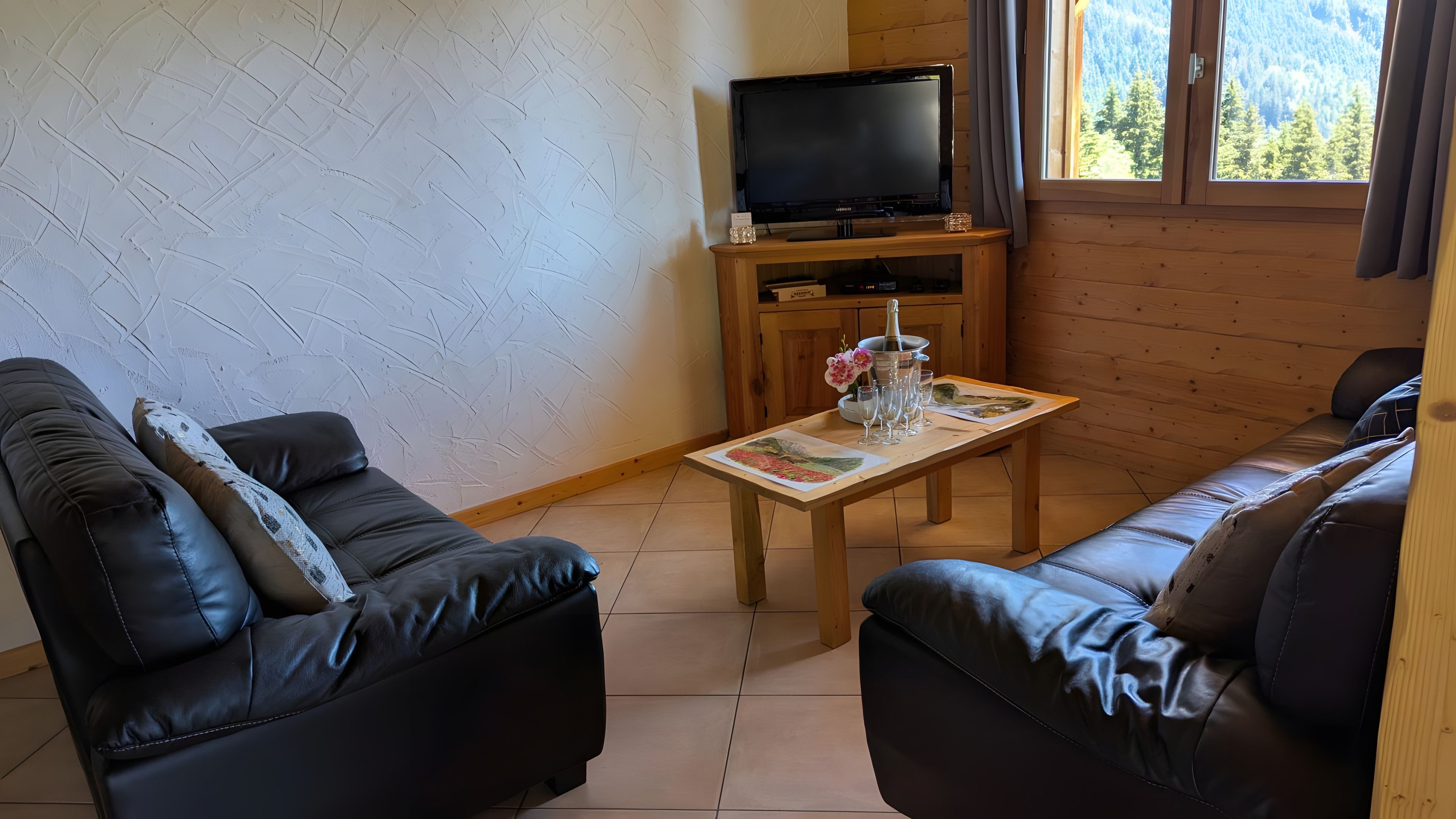 Ein möblierter Raum mit zwei Ledersofas, einem Holztisch mit einem Gemälde und Gläsern, einem Fernseher auf einem Holzständer und einem Fenster mit Blick auf die Berge.