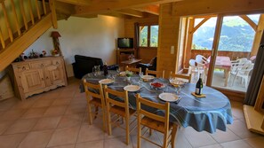 Ein Essbereich mit einem Tisch für vier Personen, Holzstühlen, einem Fernseher und Bergblick durch große Fenster.