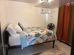 A single bed with a floral bedspread, a standing fan, and a wall-mounted air conditioner. - The Ogle Hub 1 BDR Apartment Near to Malls with AC (Georgetown)
